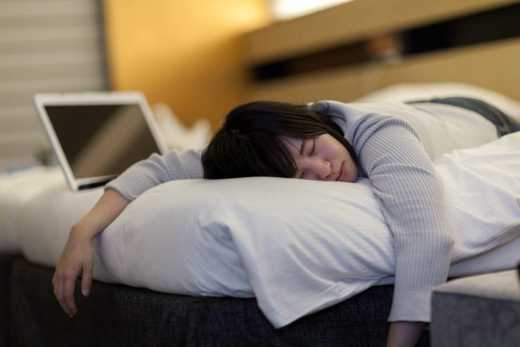 A young Japanese has fallen asleep on the bed with a laptop next to her.