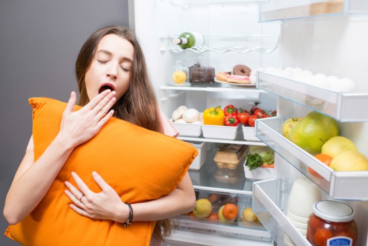 Young woman with pillow yawning standing in front of the refrigerator. Concept of eating during the night